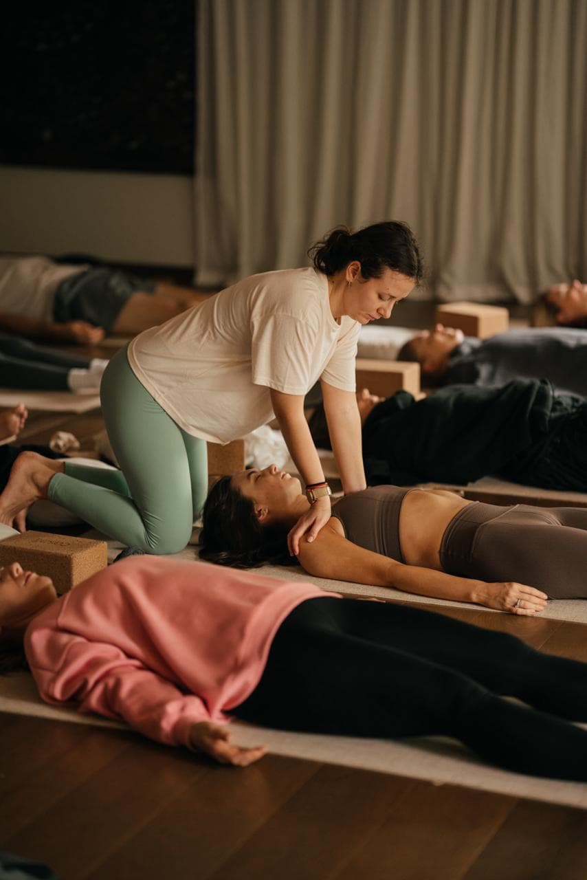 Instructor gently adjusting a student during a group relaxation pose at Mindloom, mats spread across a warm-lit studio